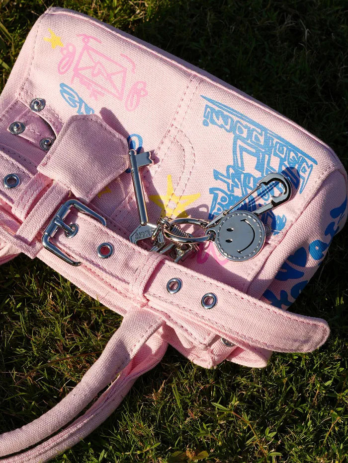 A pink canvas shoulder bag with colorful graffiti-style drawings and blue text, resting on green grass. It features silver-tone metal buckles, grommets, and a keychain with a smiley face charm attached.