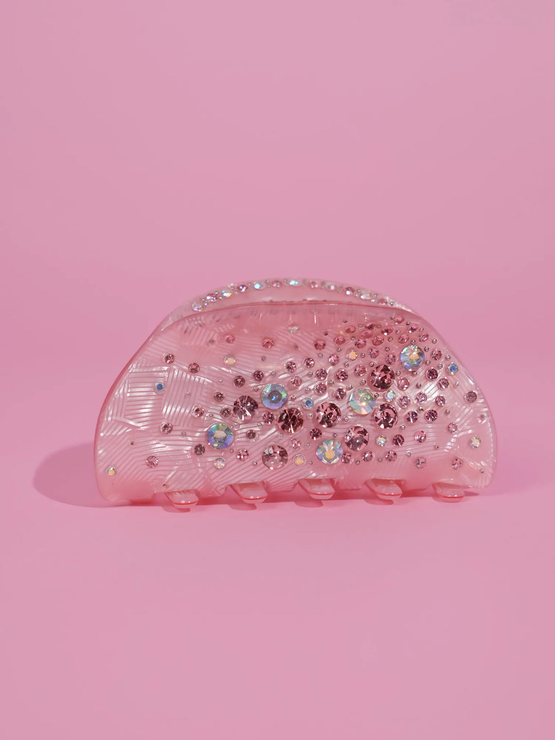A translucent pink hair claw clip decorated with iridescent rhinestones, set against a solid pink background.
